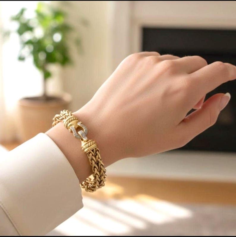 May include: A gold-toned bracelet with a braided design and a decorative clasp. The clasp features a horseshoe shape with small, clear stones. The bracelet is worn on a wrist, with a white shirt sleeve visible.