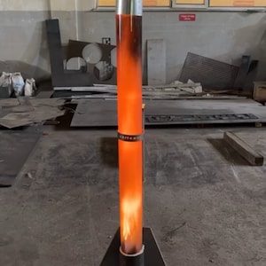 May include: A metal stand with a triangular base supports a tall, cylindrical metal tube. The tube is filled with a glowing orange liquid and a flame is visible at the top.