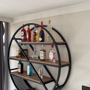 May include: A black metal circular wall shelf with wooden shelves, featuring a variety of liquor bottles, including gin, vodka, whiskey, and wine. The shelf has a wire mesh bottom for storing wine bottles.