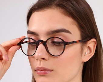 May include: A woman wearing round tortoiseshell glasses with clear lenses. The glasses have a brown frame with a tortoiseshell pattern.