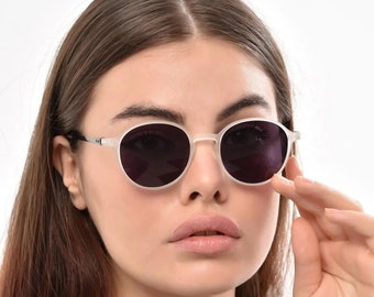 May include: A woman wearing white framed round sunglasses with dark lenses. She is wearing a black t-shirt.