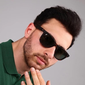 May include: A man wearing a green polo shirt and tortoiseshell sunglasses. He is looking at the camera and has his hand on his chin.