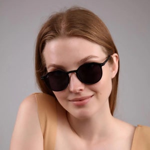 May include: A woman wearing black round sunglasses with a tan sleeveless top.