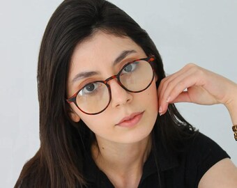 May include: A woman with long dark hair wearing round tortoiseshell glasses and a black collared shirt.