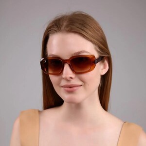 May include: A woman wearing brown square-shaped sunglasses with a brown frame. She is wearing a light brown sleeveless top.