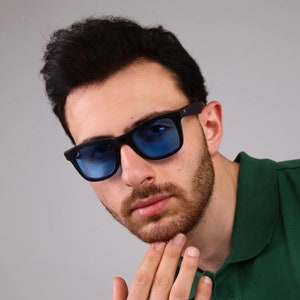 May include: A man wearing black sunglasses with blue lenses and a green polo shirt. He is looking at the camera and has his hand on his chin.