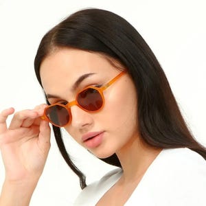 May include: A woman wearing a white shirt and orange round sunglasses. The sunglasses have a brown lens.