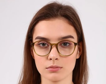 May include: A woman with long brown hair wearing round, green-framed glasses. She is wearing a white, square-neck shirt.
