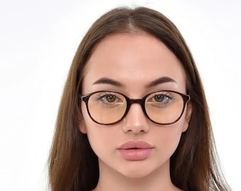 Könnte beinhalten: Eine Frau mit langen braunen Haaren trägt eine braune runde Brille mit bernsteinfarbenen Gläsern. Sie trägt ein weißes T-Shirt mit V-Ausschnitt.