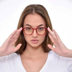 May include: A woman wearing red round eyeglasses with clear lenses. She is wearing a white v-neck t-shirt.