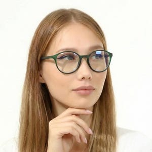 May include: Dark green, round-framed spectacles. The glasses are worn by a person with long, light brown hair and a white top. The background is plain white.