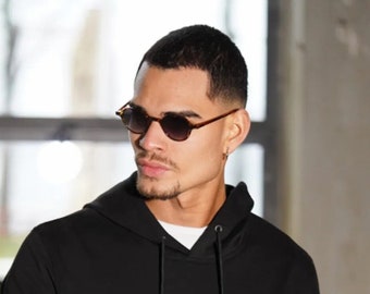 May include: A man wearing a black hooded sweatshirt and round, dark brown sunglasses.