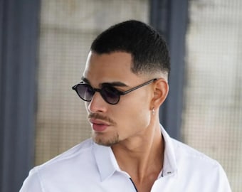 Könnte beinhalten: Ein Mann mit kurzen, dunklen Haaren, der ein weißes Hemd mit Knöpfen und eine schwarze runde Sonnenbrille trägt.