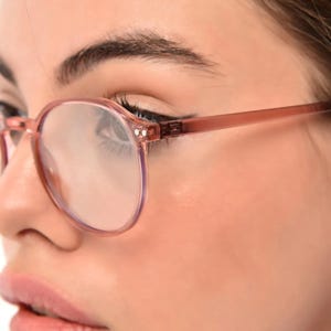 May include: A close-up of a person wearing pink round glasses with clear lenses. The glasses have a thin, delicate frame.