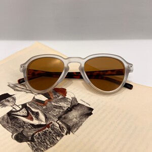 May include: A pair of frosted white sunglasses with brown lenses. The sunglasses are sitting on a book with a black and white illustration.