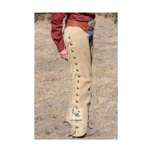 Puede incluir: Chaps de cuero de gamuza color beige con flecos a lo largo de la costura exterior y botones dorados decorativos. Los chaps se usan con un cinturón de cuero marrón y una hebilla plateada. La imagen muestra un primer plano de los chaps.