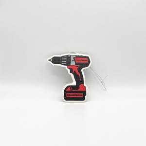 K&ouml;nnte beinhalten: Ein schwarzer, roter und wei&szlig;er Lufterfrischer in Form eines Akkubohrers. Der Lufterfrischer tr&auml;gt unten den Text "TRADEFRESHUK" und oben "TP TRADFRESHUK".