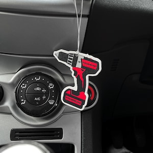 K&ouml;nnte beinhalten: Ein schwarz-rot-wei&szlig;er Lufterfrischer in Form eines Elektrowerkzeugs h&auml;ngt im Auto. Der Lufterfrischer tr&auml;gt den Text "#TRADEFRESHUK" und "TP". Das Armaturenbrett des Autos ist grau.