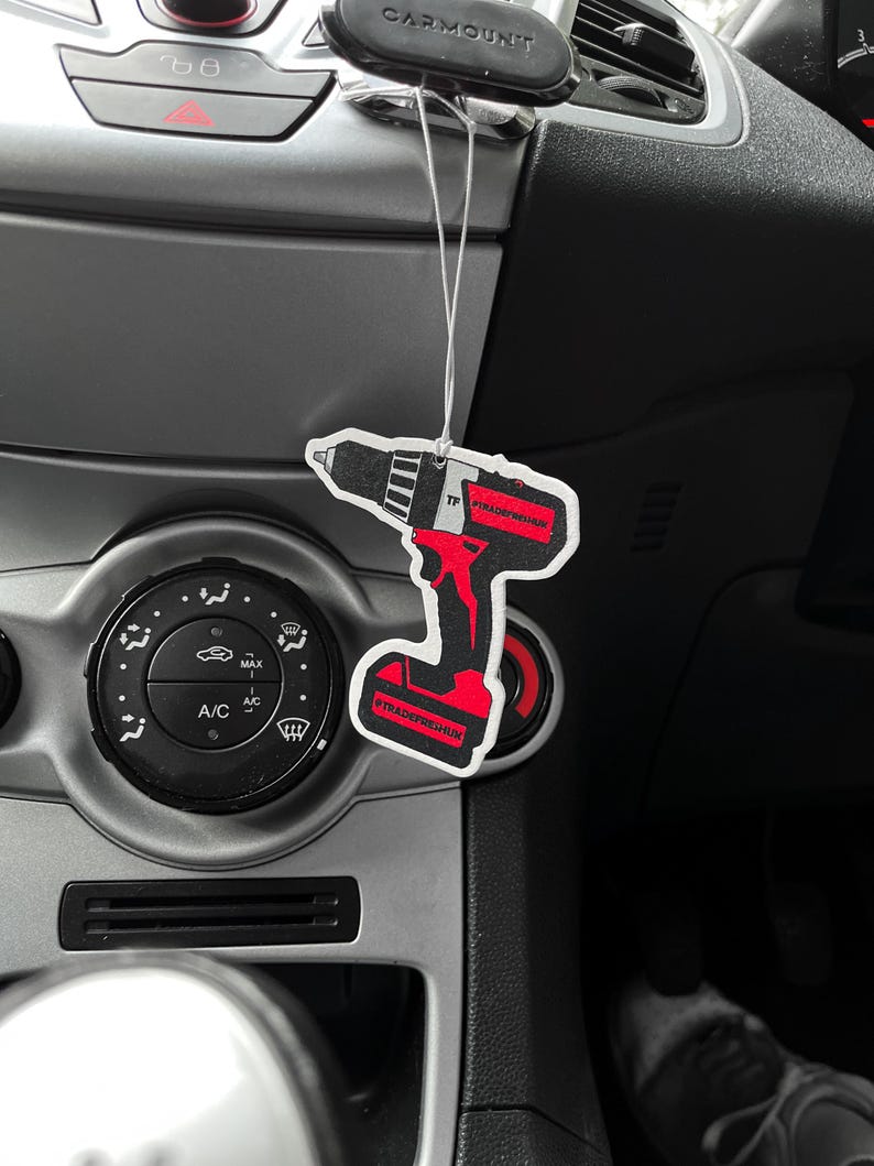 K&ouml;nnte beinhalten: Ein schwarz-rot-wei&szlig;er Lufterfrischer in Form eines Elektrowerkzeugs h&auml;ngt im Auto. Der Lufterfrischer tr&auml;gt den Text "#TRADEFRESHUK" und "TP". Das Armaturenbrett des Autos ist grau.