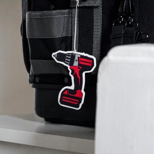 K&ouml;nnte beinhalten: Eine schwarze und graue Werkzeugtasche mit grauen Riemen und schwarzer Hardware. Ein rot-schwarzer, bohrerf&ouml;rmiger Lufterfrischer h&auml;ngt an der Tasche. Der Lufterfrischer tr&auml;gt den Text "TRADERFRESHUK".