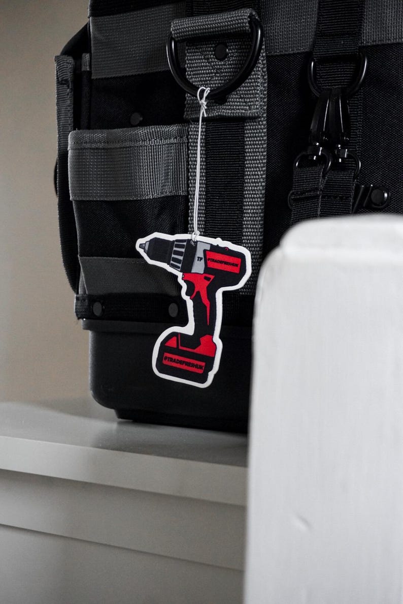 K&ouml;nnte beinhalten: Eine schwarze und graue Werkzeugtasche mit grauen Riemen und schwarzer Hardware. Ein rot-schwarzer, bohrerf&ouml;rmiger Lufterfrischer h&auml;ngt an der Tasche. Der Lufterfrischer tr&auml;gt den Text "TRADERFRESHUK".
