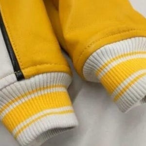 May include: Close-up of a yellow and white varsity jacket. The jacket features yellow sleeves and a white body with a black stripe. The cuffs are white with yellow stripes.