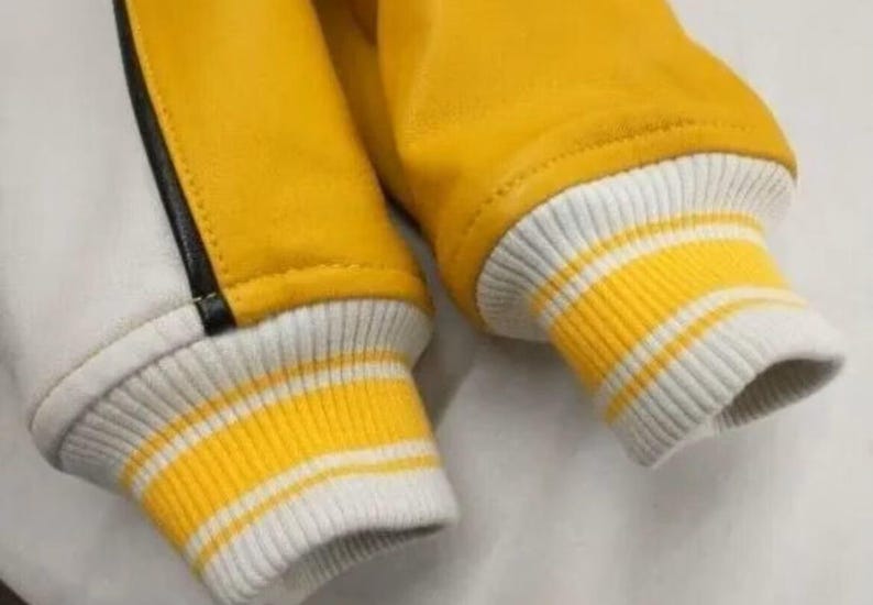 May include: Close-up of a yellow and white varsity jacket. The jacket features yellow sleeves and a white body with a black stripe. The cuffs are white with yellow stripes.