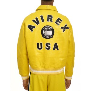 Puede incluir: Chaqueta de cuero amarilla con la palabra "AVIREX" y "USA" en la espalda. La chaqueta tambi&eacute;n muestra "ALL AMERICANS ESTABLISHED 1975". Tiene un acabado texturizado y ribetes a rayas blancas y amarillas.