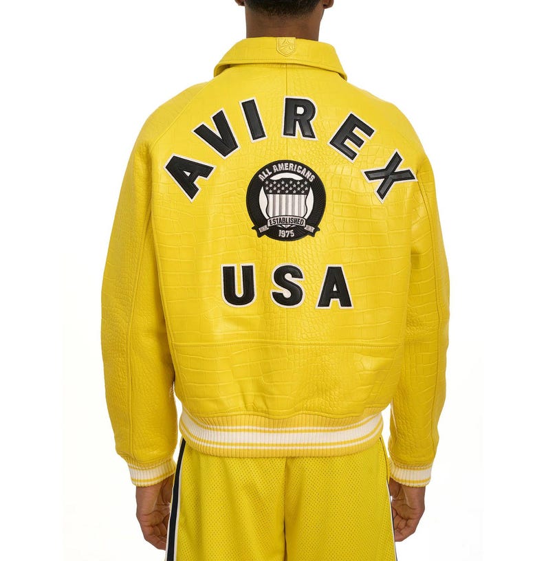 Puede incluir: Chaqueta de cuero amarilla con la palabra "AVIREX" y "USA" en la espalda. La chaqueta tambi&eacute;n muestra "ALL AMERICANS ESTABLISHED 1975". Tiene un acabado texturizado y ribetes a rayas blancas y amarillas.