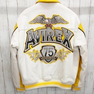 May include: White leather jacket with yellow detailing, featuring the word "AVIREX" in large lettering on the back. An eagle emblem and the text "75 AVIATOR'S KING" are also visible.