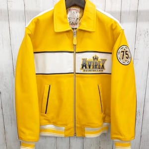 May include: A bright yellow leather jacket with white stripes and trim. The jacket displays the "AVIREX" logo and a "75" patch. It has a zip closure, collar, and ribbed cuffs and hem. The jacket is suspended on a wooden hanger.