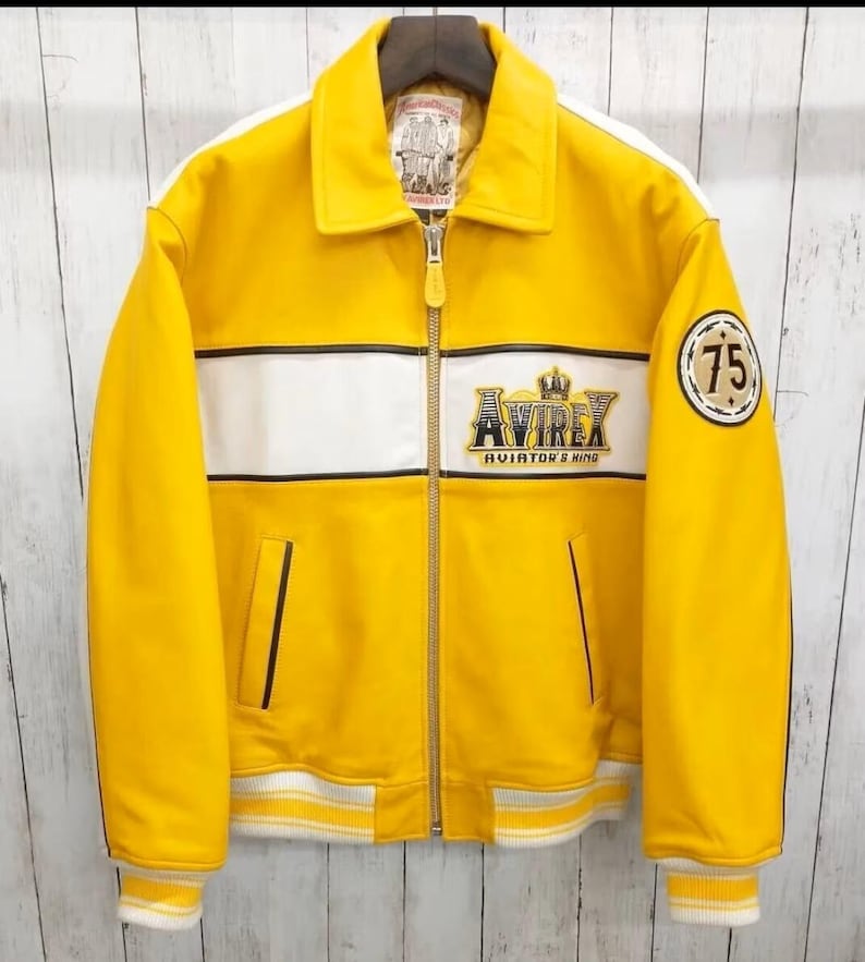 May include: A bright yellow leather jacket with white stripes and trim. The jacket displays the "AVIREX" logo and a "75" patch. It has a zip closure, collar, and ribbed cuffs and hem. The jacket is suspended on a wooden hanger.