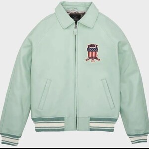 May include: A light green leather jacket with a collared neckline and a zipper closure. The jacket features a patch on the chest with a red, white, and blue emblem and the word "AVIREX". The cuffs and hem have a striped design.