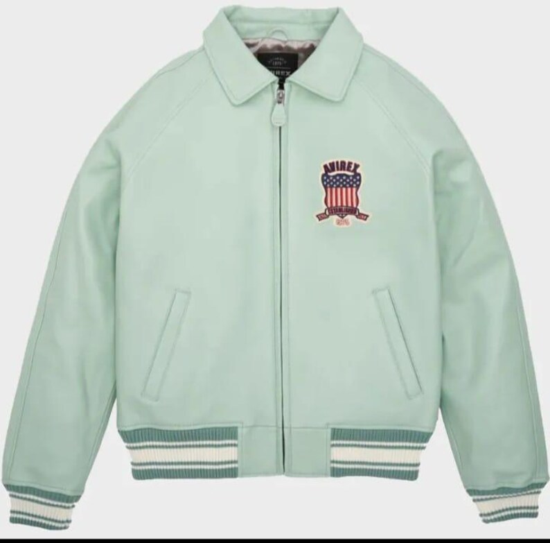 May include: A light green leather jacket with a collared neckline and a zipper closure. The jacket features a patch on the chest with a red, white, and blue emblem and the word "AVIREX". The cuffs and hem have a striped design.