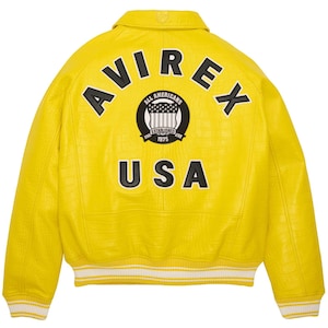 Puede incluir: Chaqueta de cuero amarillo brillante con las palabras "AVIREX" y "USA" en letras negras. Un parche circular con "ALL AMERICANS ESTABLISHED 1975" est&aacute; centrado encima de "USA". La chaqueta tiene un patr&oacute;n texturizado y ribetes blancos.