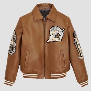 May include: A brown leather jacket with a collared neckline and a zip-up front. The jacket features white and beige embroidered patches, including a tiger design and text on the sleeves. The cuffs and hem have white stripes.