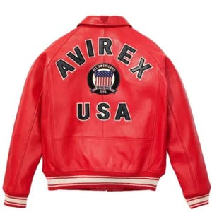 Pode incluir: Casaco de couro vermelho com a palavra "AVIREX" arqueada sobre os ombros e "USA" abaixo. O casaco apresenta uma faixa listrada branca e vermelha nos punhos e na bainha. Um patch circular com as palavras "ALL AMERICANS" e uma bandeira dos EUA está centrado nas costas.
