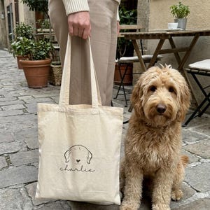 Könnte beinhalten: Eine beigefarbene Tragetasche aus Segeltuch mit einer schwarzen Strichzeichnung eines Hundekopfes mit einer kleinen herzförmigen Nase und dem Namen "charlie" in kursiver Schrift, der unter der Grafik auf der Vorderseite zentriert ist.