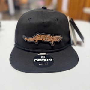 May include: Black baseball cap with a brown alligator patch. The cap has a curved brim, a black rope detail, and the brand name "DECKY" on a sticker. The alligator patch is detailed with brown spots.