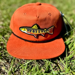 May include: A burnt orange baseball cap with a black brim and a brown trout fish patch on the front.
