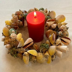 Peut inclure: Une bougie rouge brûlant au centre d'une couronne faite de coquillages. Les coquillages sont de différentes couleurs et formes, notamment blanc, brun, jaune et vert.