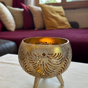 May include: A gold metal candle holder with a decorative cut-out design and three legs. The candle holder is sitting on a white table cloth.