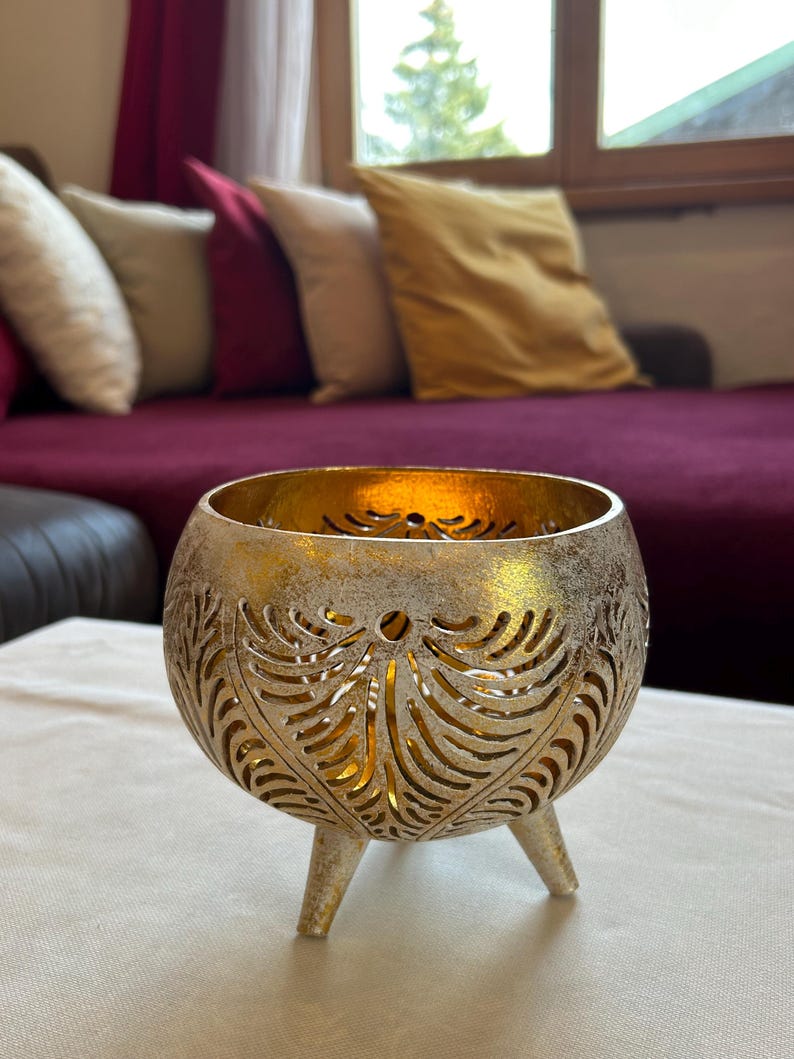 May include: A gold metal candle holder with a decorative cut-out design and three legs. The candle holder is sitting on a white table cloth.
