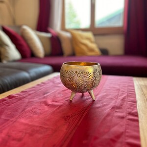 May include: A gold metal candle holder with an intricate design sits on a red table runner. The candle holder has three legs and is made of a delicate, openwork material.