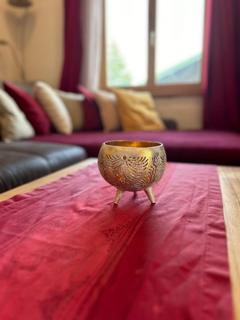 May include: A gold metal candle holder with an intricate design sits on a red table runner. The candle holder has three legs and is made of a delicate, openwork material.