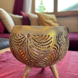 May include: A gold metal candle holder with a decorative, cut-out design. The holder has three legs and a round, open top.