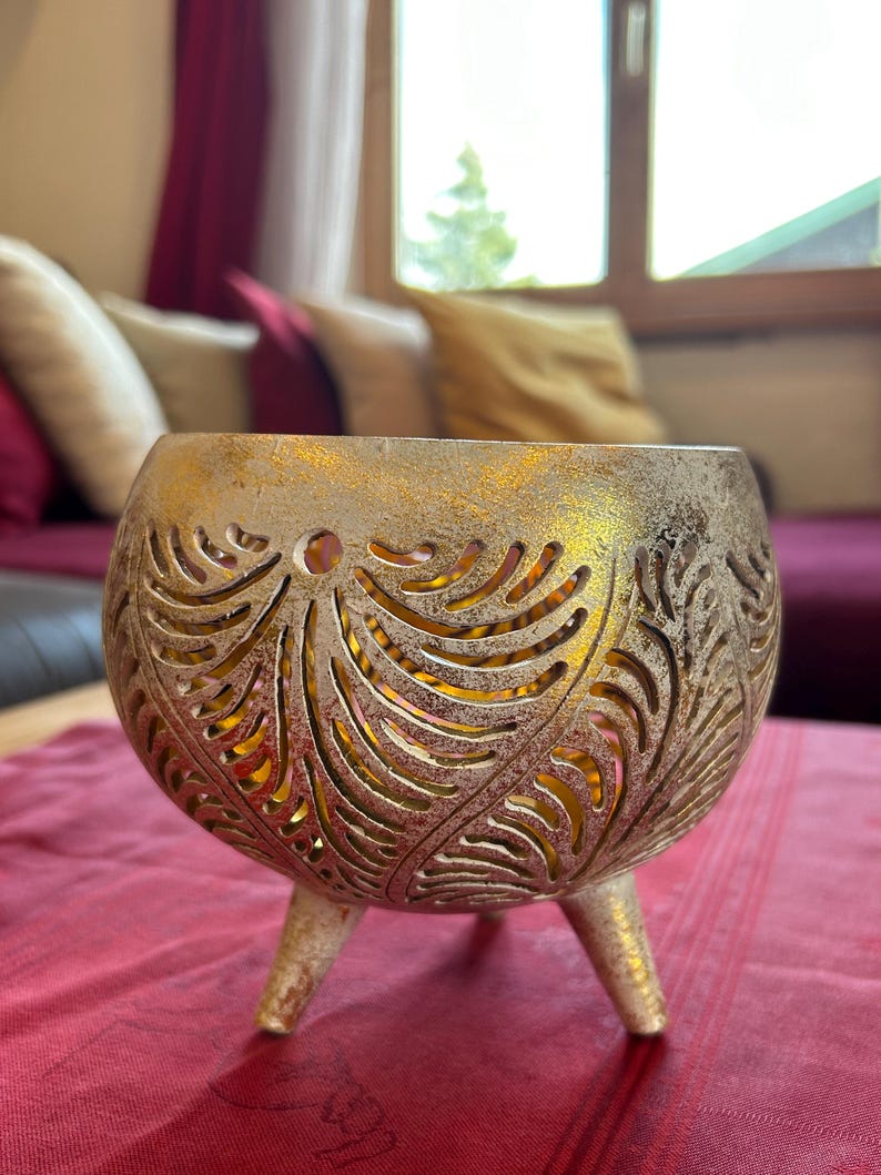 May include: A gold metal candle holder with a decorative, cut-out design. The holder has three legs and a round, open top.