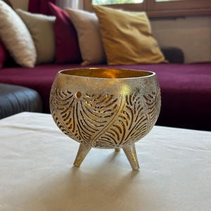 May include: A gold and silver metal candle holder with a decorative leaf pattern. The holder has three legs and is sitting on a white surface.