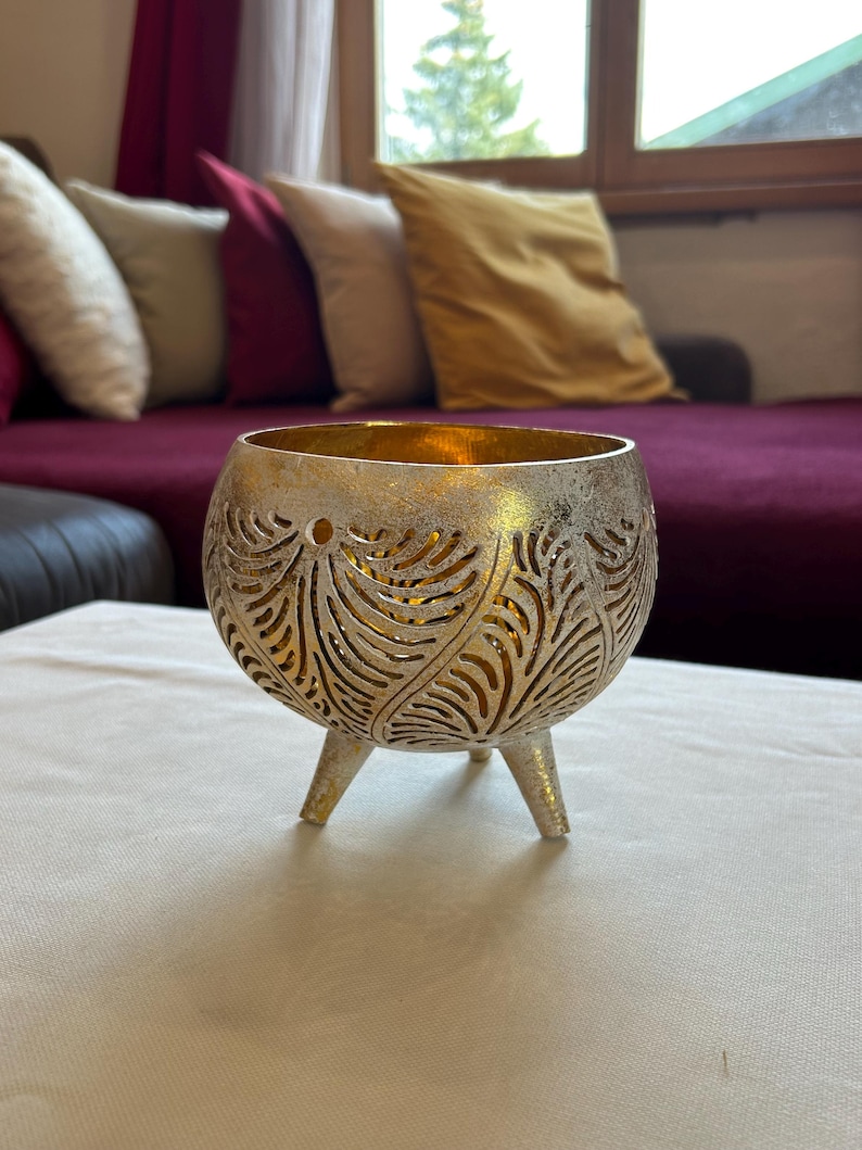 May include: A gold and silver metal candle holder with a decorative leaf pattern. The holder has three legs and is sitting on a white surface.