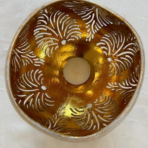 May include: A gold-painted coconut shell candle holder with a white, intricate, carved design. The holder has a small, round opening in the center for a tea light candle.